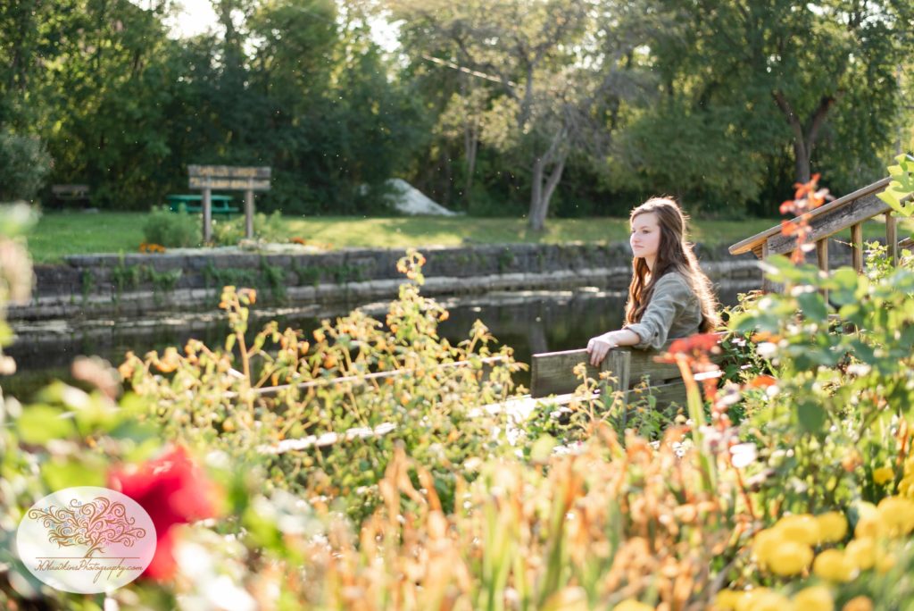 High school senior girl sits on a bench on the dock at the historic Sims store of the Erie Canal Park in Camillus CNY with flowers in the foreground