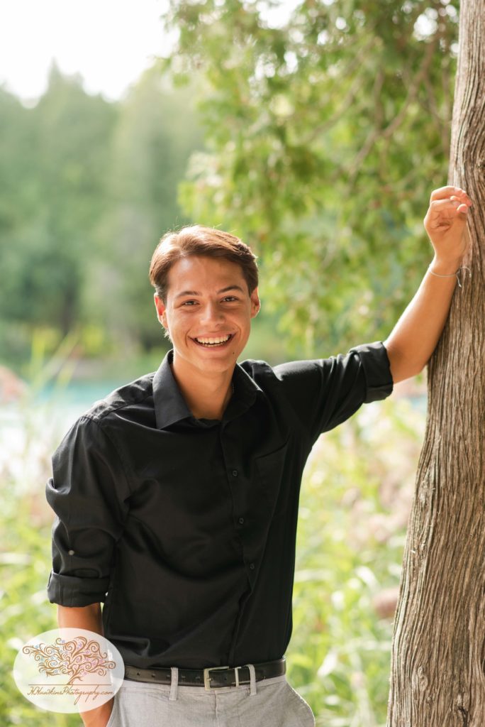 high school senior boy in a black shirt leans against a tree in front of Green Lakes State Park with his hands up on the tree