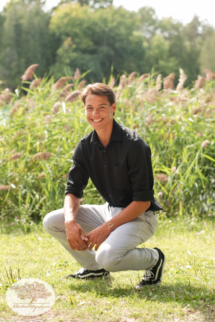 high school senior boy in a black shirt squats in front of Green Lakes State Park