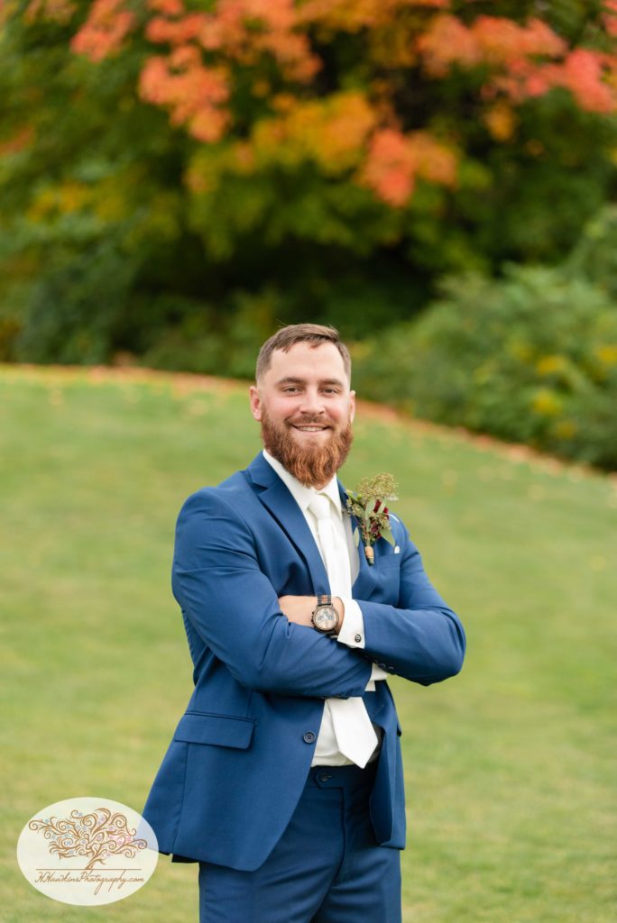 Groom smiles for a wedding portrait in his blue suit jacket with arms crossed over his chest at The View at Sunset Pines Executive Golf Course