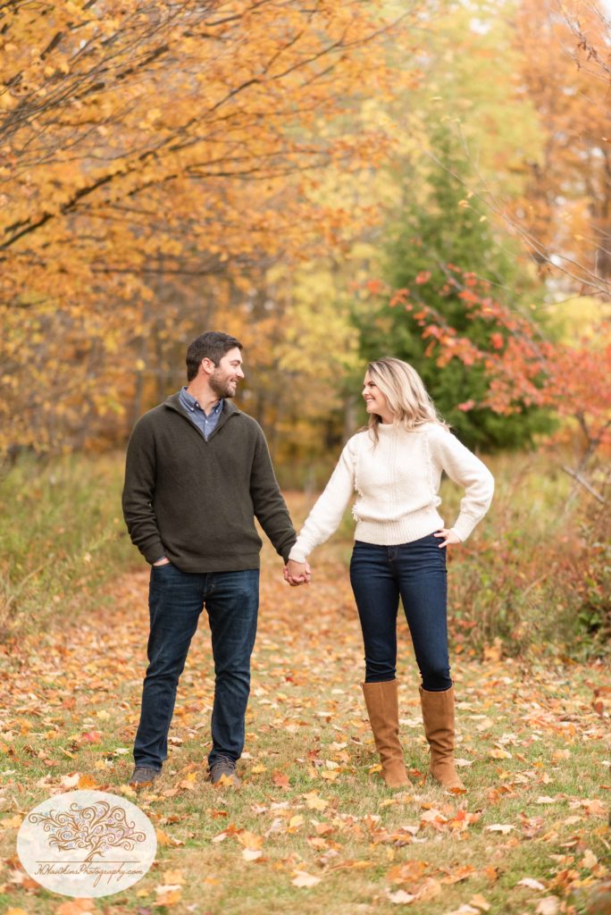 Engaged couple stands holding hands and looking at each other with all the fall foliage around them at Baltimore Woods Marcellus NY