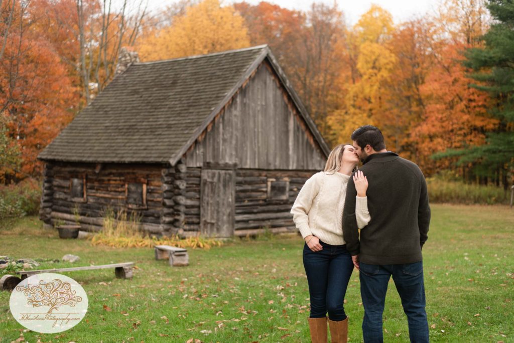 Bride holds groom's arm as she kisses him in front of the log cabin at Baltimore Woods Marcellus NY