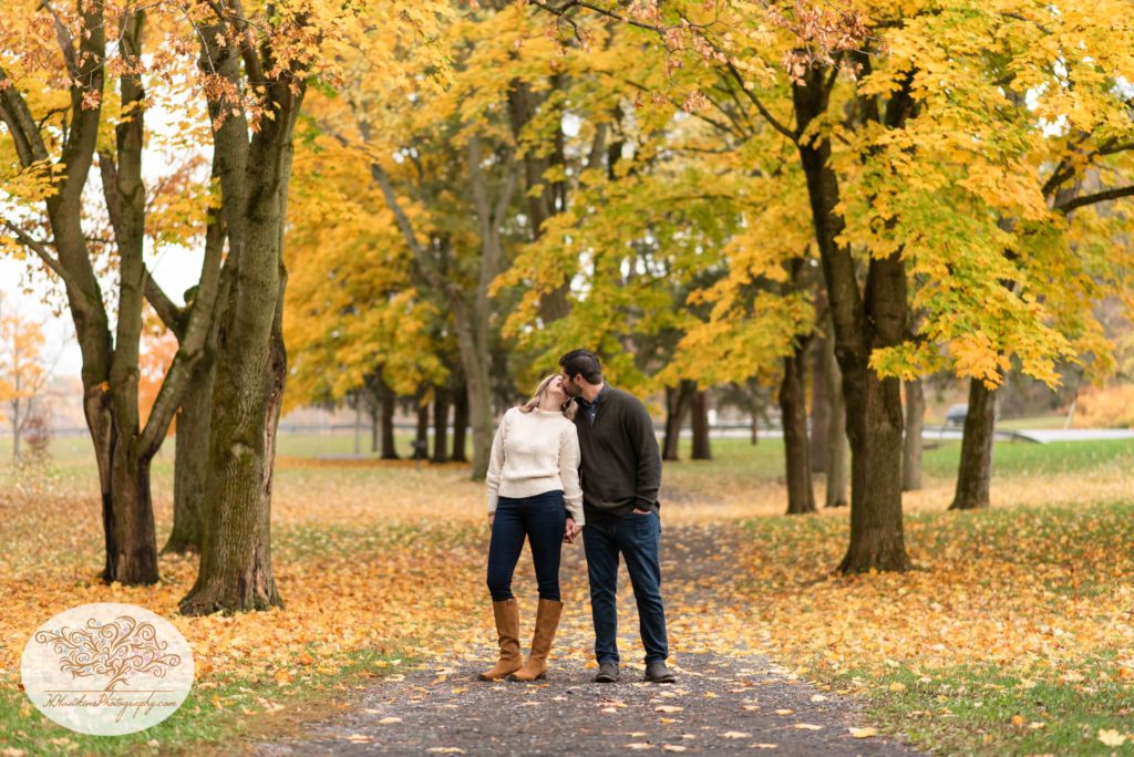 Bride and groom to be kiss on a path through yellow autumn trees at Marcellus Park for their Syracuse fall engagement pictures