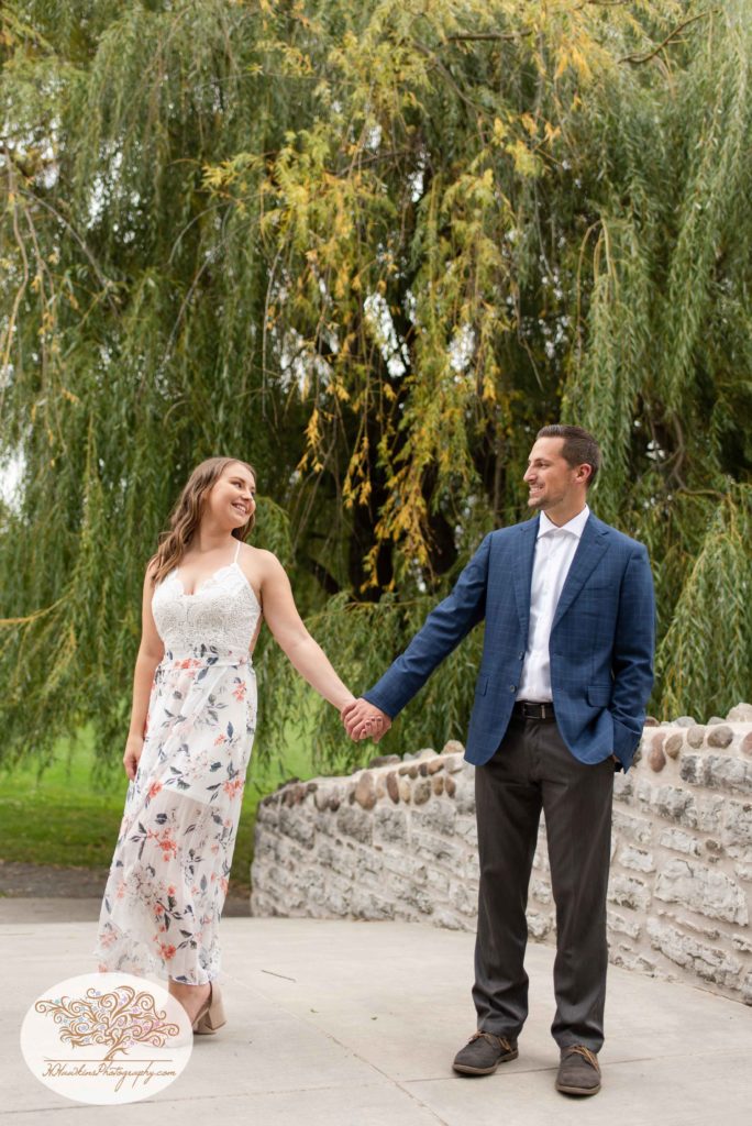 Bride in a floral dress and groom in his suit jacket stand on stone bridge at Upper Onondaga Park in Syracuse NY during their engagement session