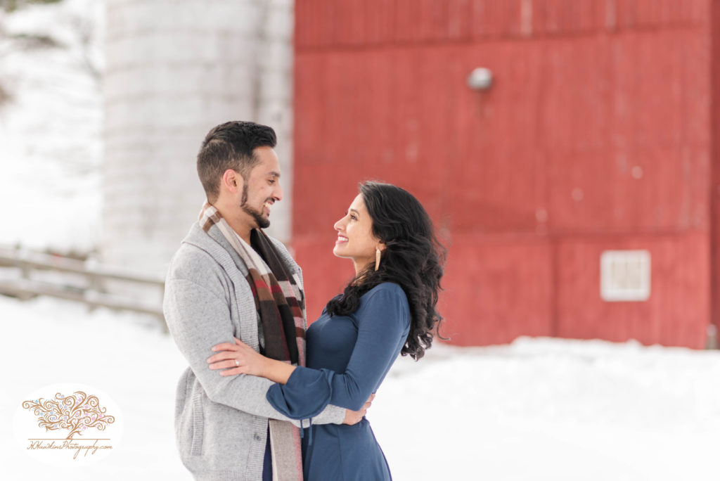 Bride and groom to be stand in front of red barn and silo at Critz Farms for their snowy engagement pictures in Cazenovia NY