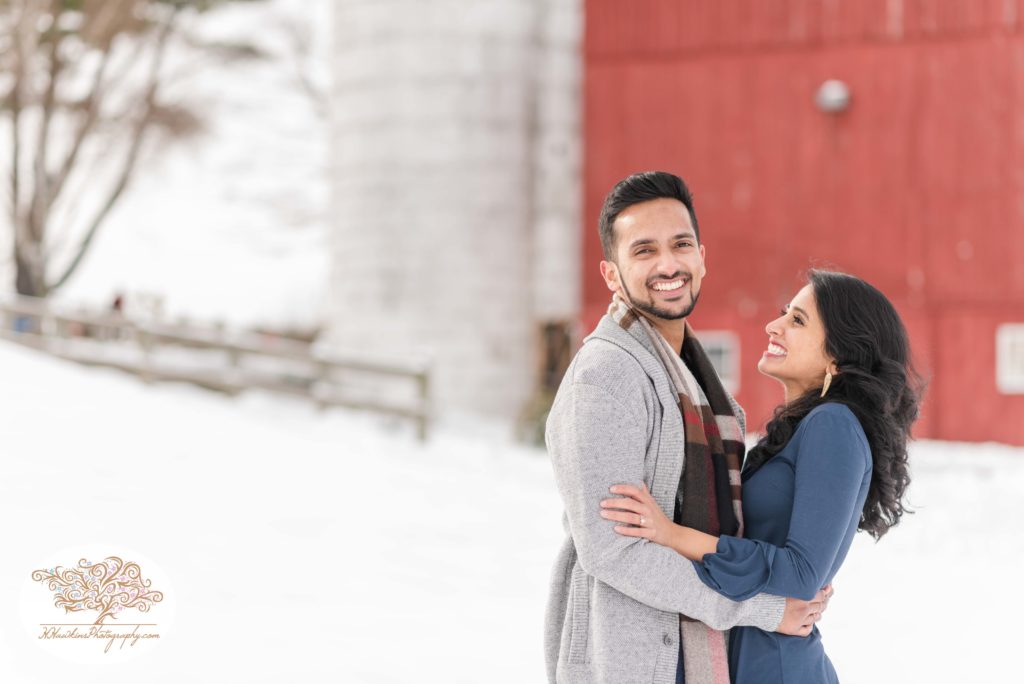 Bride and groom to be stand in front of red barn and silo at Critz Farms for their snowy engagement pictures in Cazenovia NY