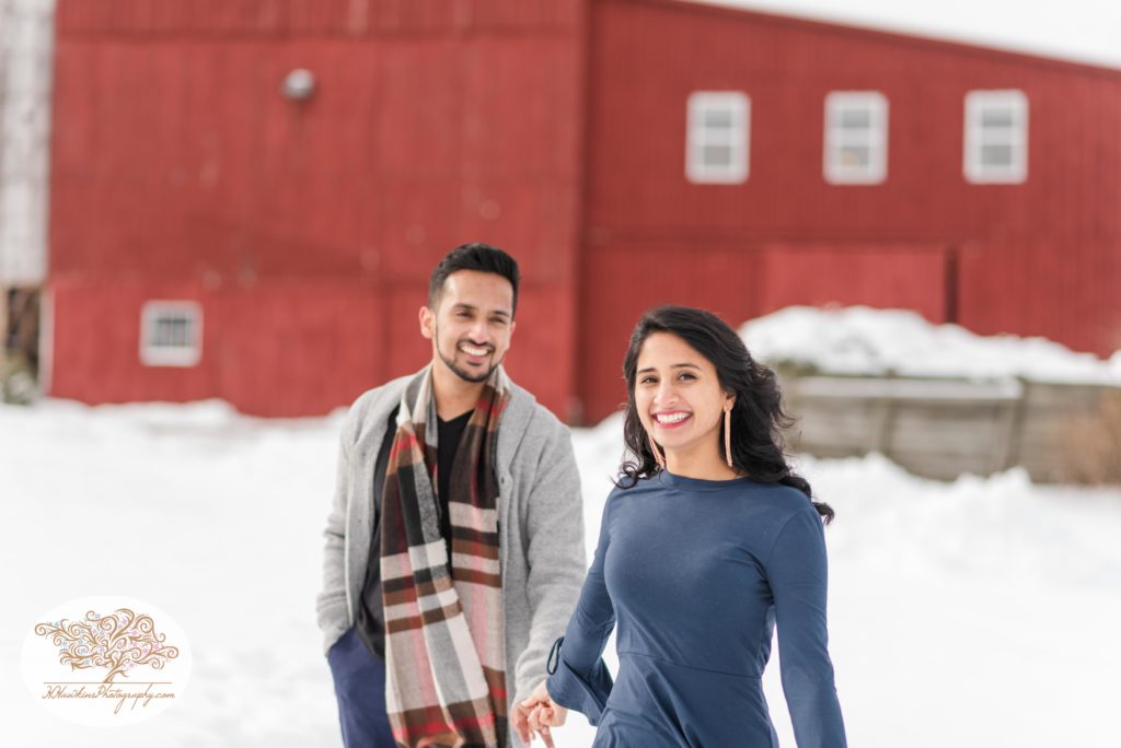 Bride looks at the camera as she holds groom's hand during their snowy engagement pictures at Critz Farms
