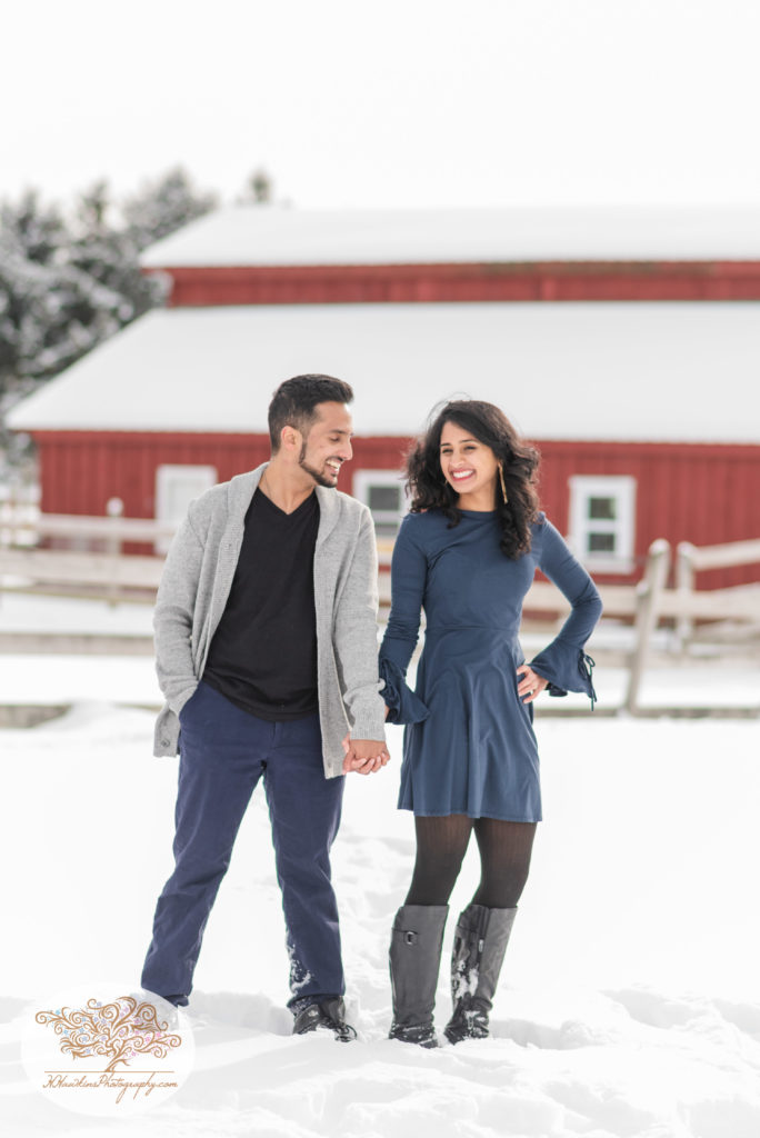Engaged couple holds hands standing in the snow in front of red barn at Critz Farms in Cazenovia NY