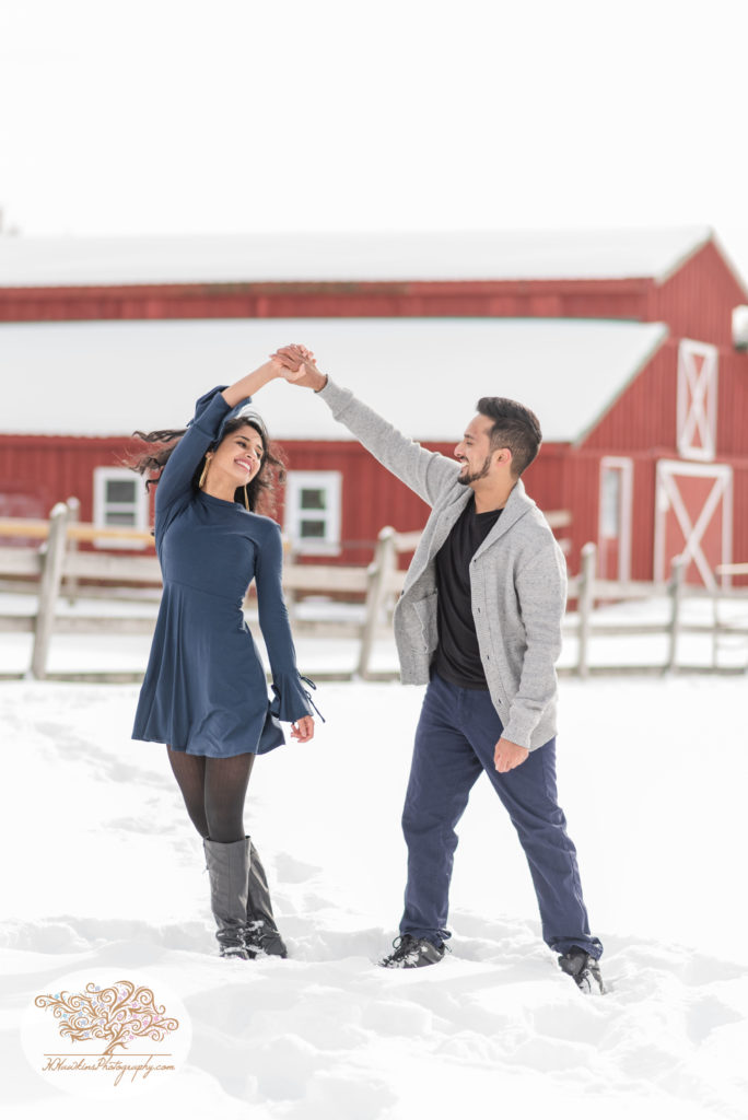 Groom spins bride to be in the snow in her blue dress in front of red barn at Critz Farms in Cazenovia NY