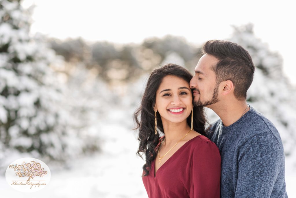 Groom kisses bride to be on the cheek as she wears a wine red dress during their engagement pictures at Critz Farms in the snow