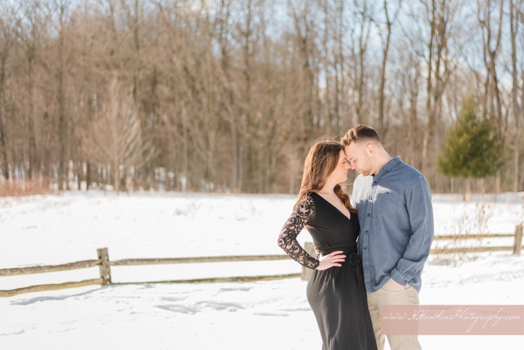Bride and groom to be stand in the snow in front of a split rail fence at Baltimore Woods in Marcellus NY for their engagement pictures