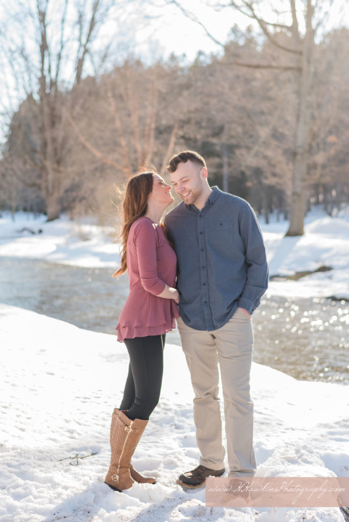Bride in pink shirt whispers secret into groom's ear as they stand in the snow in front of a river in Marcellus NY for their engagement photos