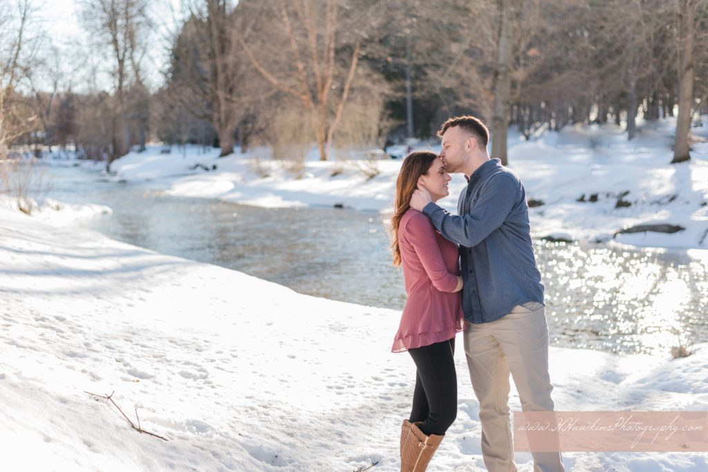 Groom kisses bride to be on the forehead during their sunny engagement pictures in the snow