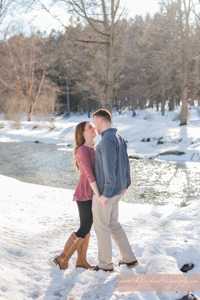 Bride and groom to be touch noses as they stand together in the snow in front of river for their engagement pictures