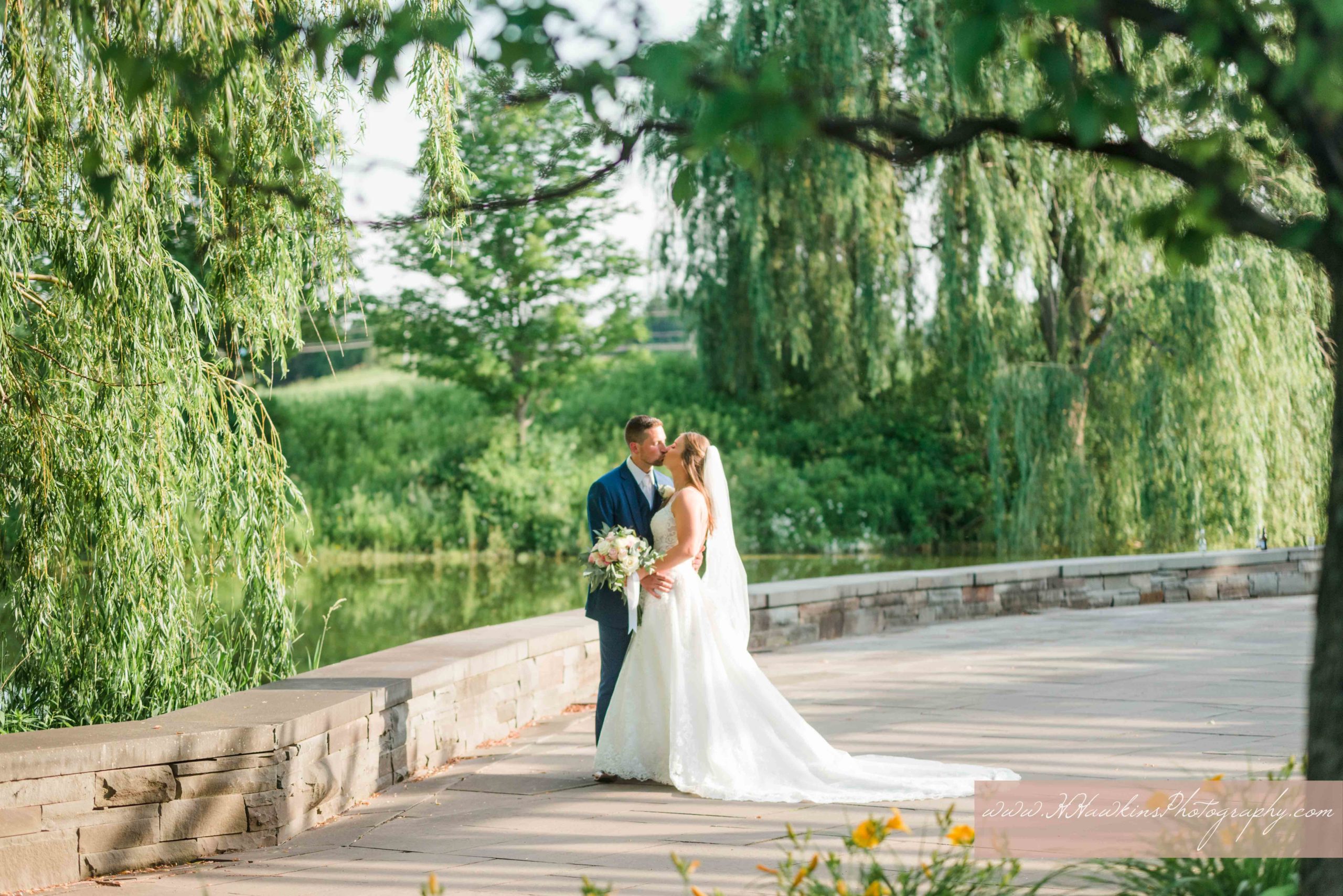 Bride and groom kiss with weeping willow trees around the at Turning Stone Lodge Great Lawn