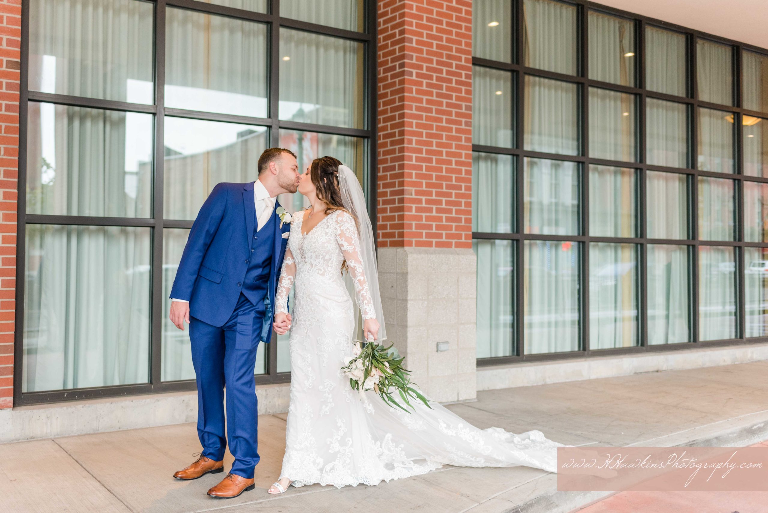 Portraits of bride in lace Kitty Chen dress and groom in navy blue suit with a brick background in the Porte-cochère of the Courtyard Marriott in Armory Square in downtown Syracuse