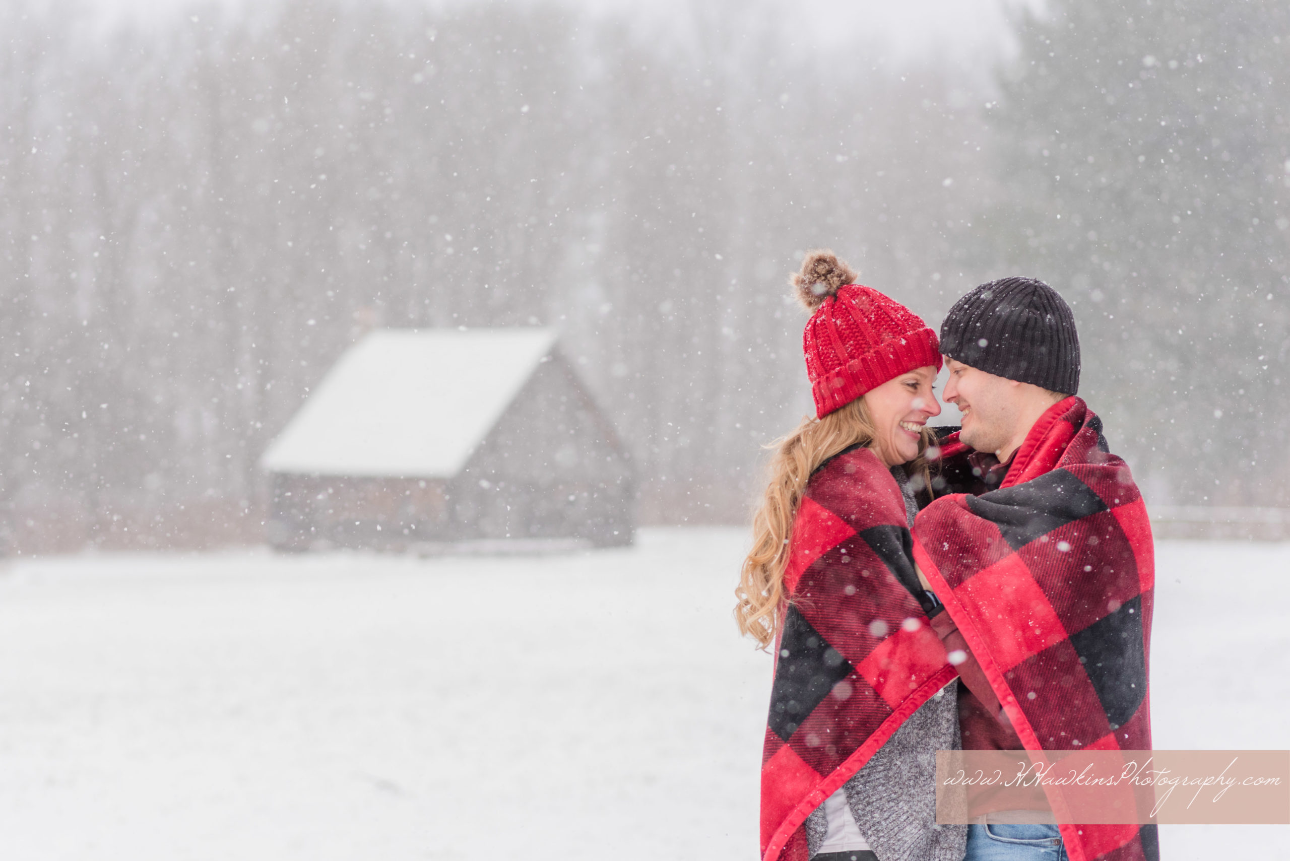 Bride and groom to be wrapped in a warm red and black plaid blanket in the snow in front of log cabin at Baltimore Woods in Marcellus NY