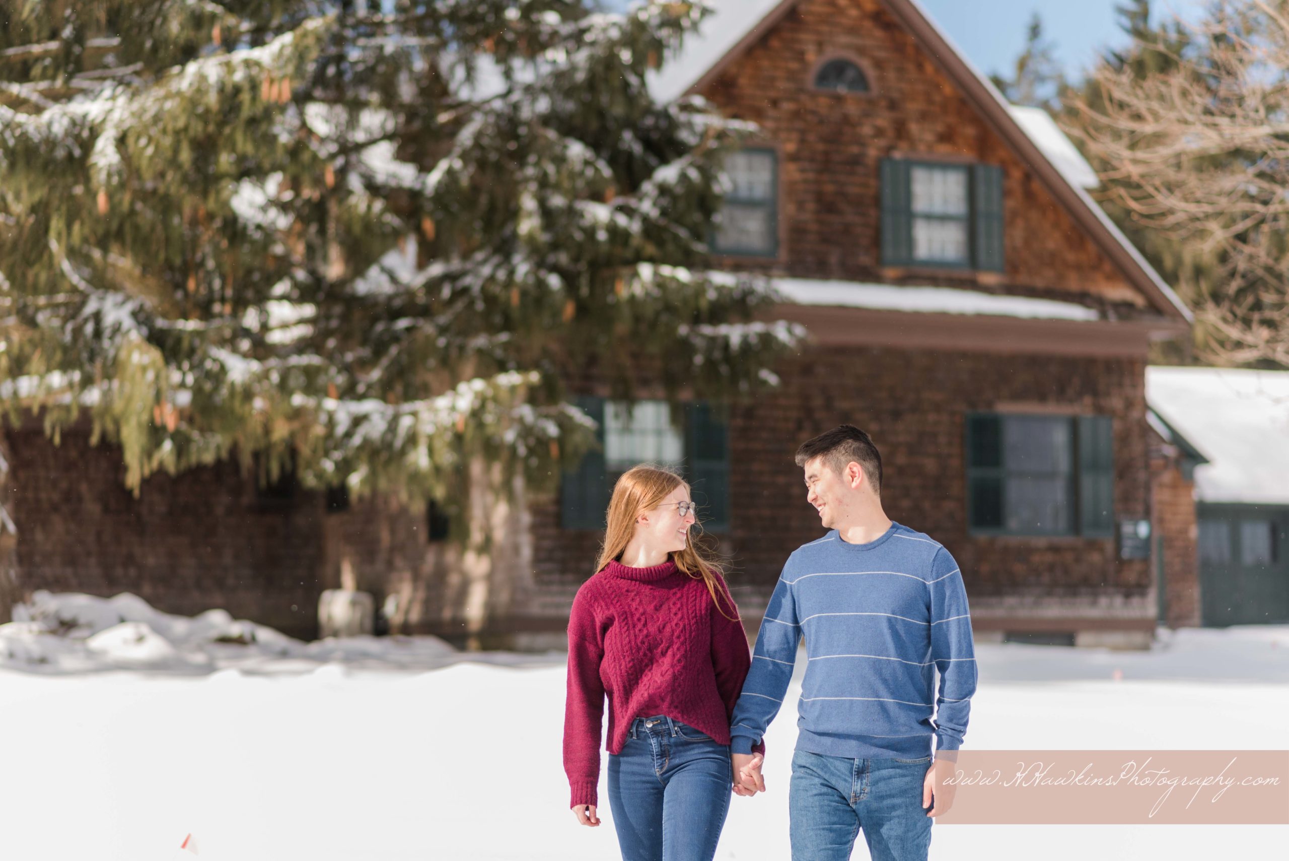 Bride and groom to be walk in front of snow covered house and pine tree at Lorenzo Historic State Park in Cazenovia NY by Syracuse photographer