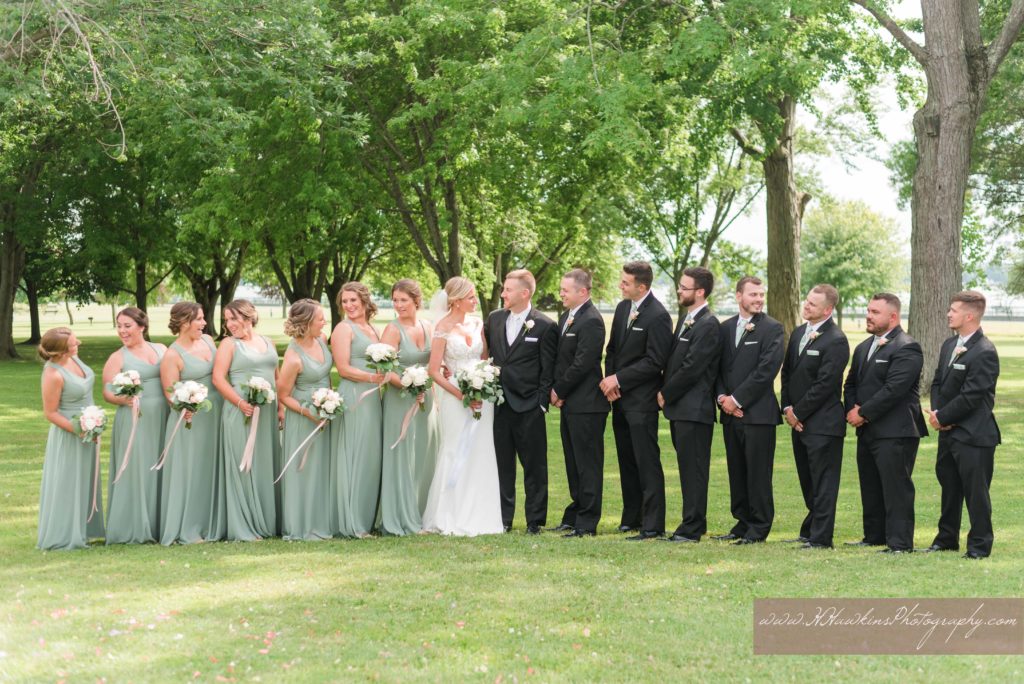 Bridal party on lawn of Emerson Park pavilion in sage green dresses and black tuxes
