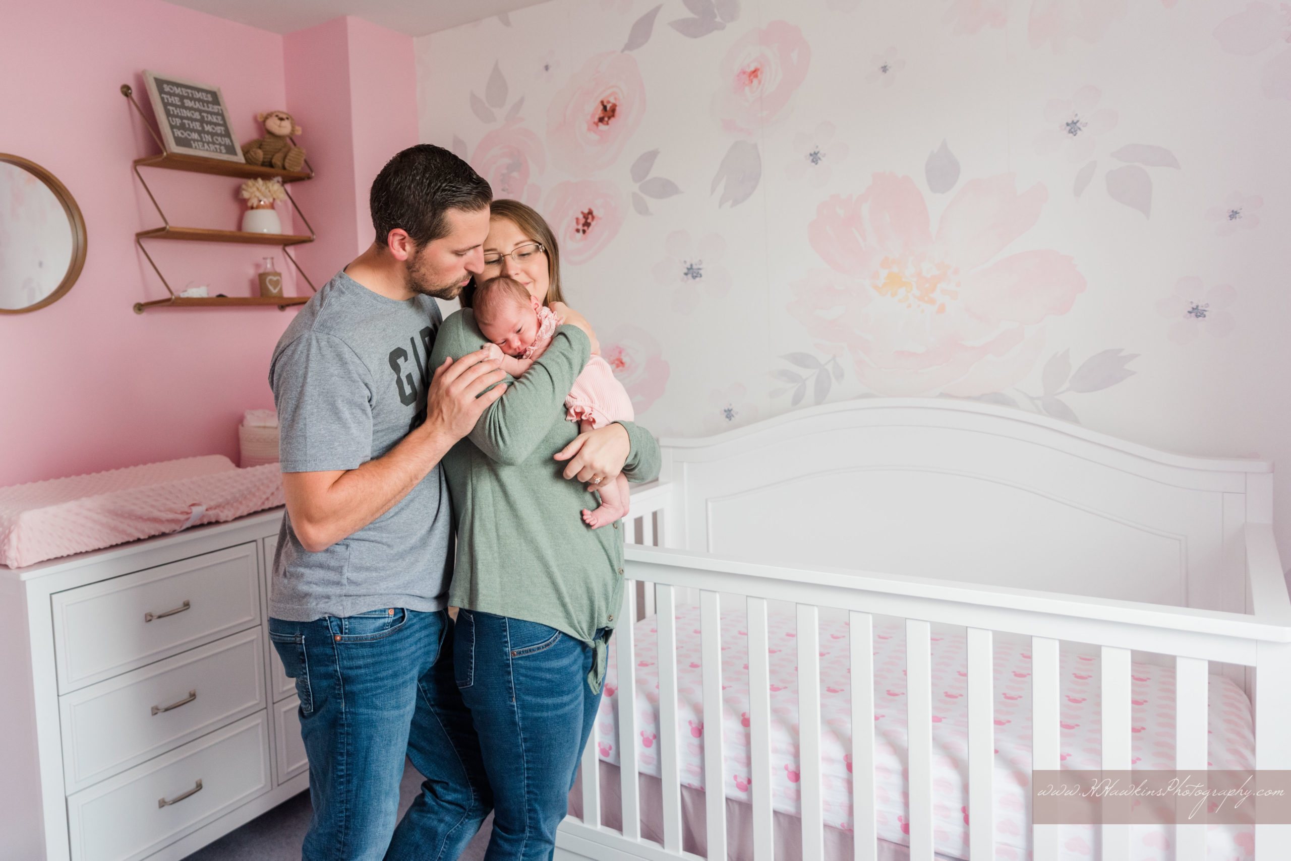 Dad kisses mom on head while mom kisses baby girl in floral pink nursery during lifestyle newborn pictures session by Orlando family photographer
