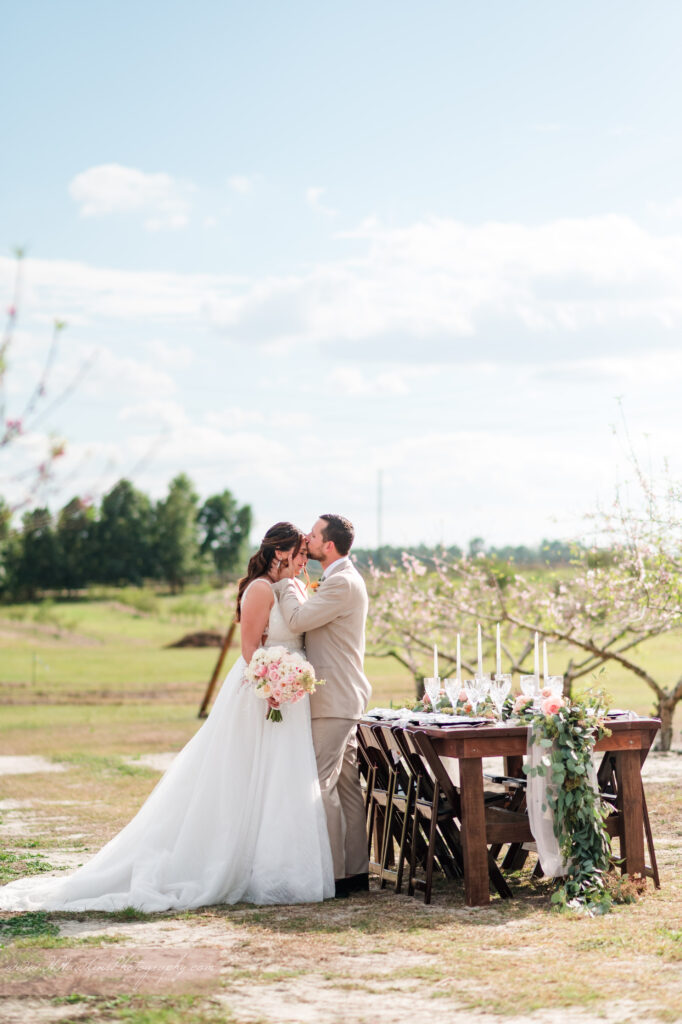 Groom kisses bride in front of wedding reception table with flowers by Lily's Flower Shop and set by Crystal Event Rentals and Design in Acres of Grace wedding venue peach blossom orchard