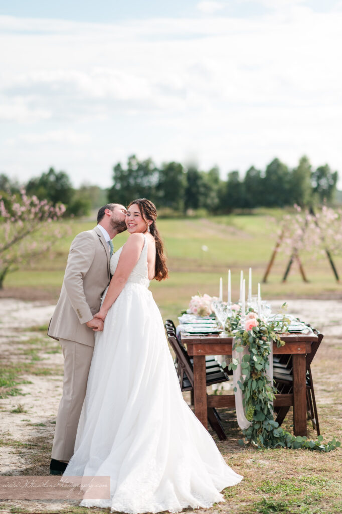Groom kisses bride in front of wedding reception table with flowers by Lily's Flower Shop and set by Crystal Event Rentals and Design in Acres of Grace wedding venue peach blossom orchard