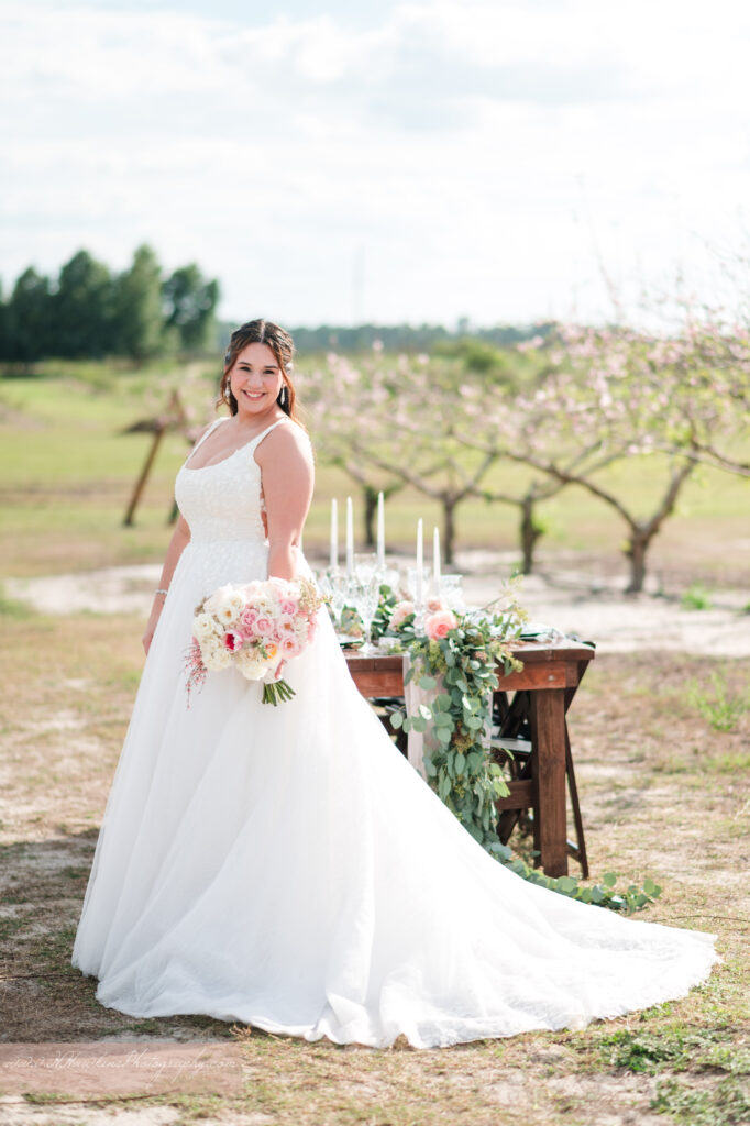 Bride and her bouquet stands in front of wedding reception table in middle of peach blossom orchard at Acres of Grace Family Farms in Howey in the Hills FL