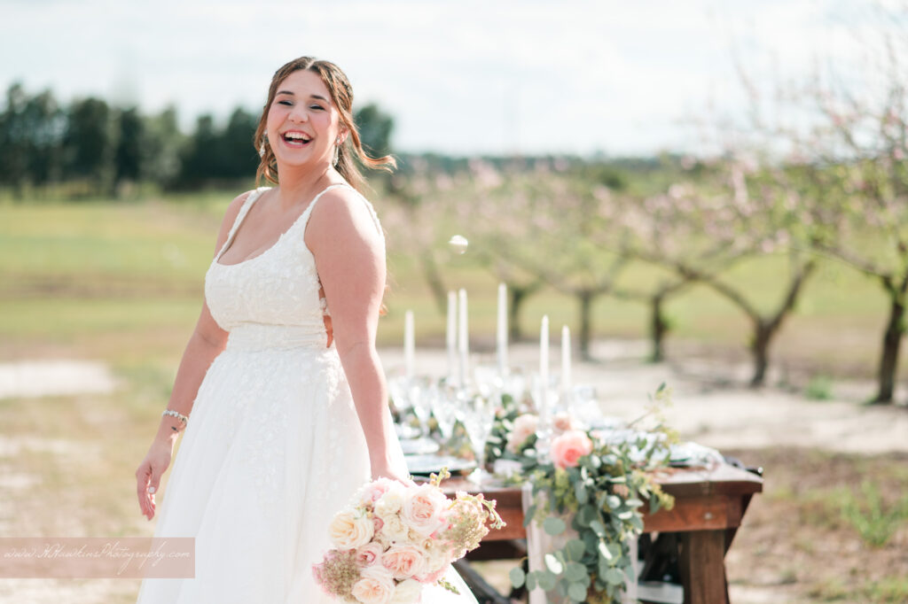 bride laughs in front of wedding reception table with flowers by Lily's Flower Shop and set by Crystal Event Rentals and Design in Acres of Grace wedding venue peach blossom orchard