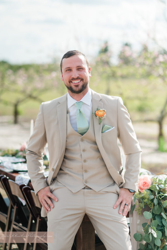 Groom in a tux rented by It Suits Me in Lake Mary sits on the end of wedding reception table in middle of peach blossom orchard at Acres of Grace Family Farms in Howey in the Hills FL