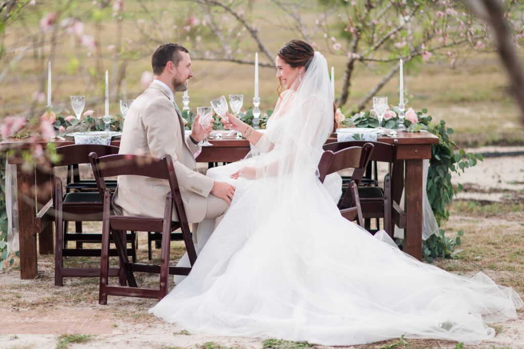 Bride and groom sit at wedding reception table in peach blossom orchard at Acres of Grace Family Farms wedding venue in Howey in the Hills
