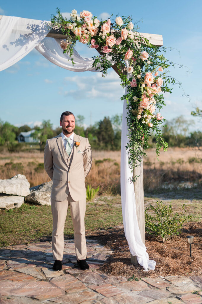 Groom awaits his bride under wedding arch decorated by pink florals and fairy tale vines by Lily's Flower Shop at Acres of Grace Family Farms wedding venue
