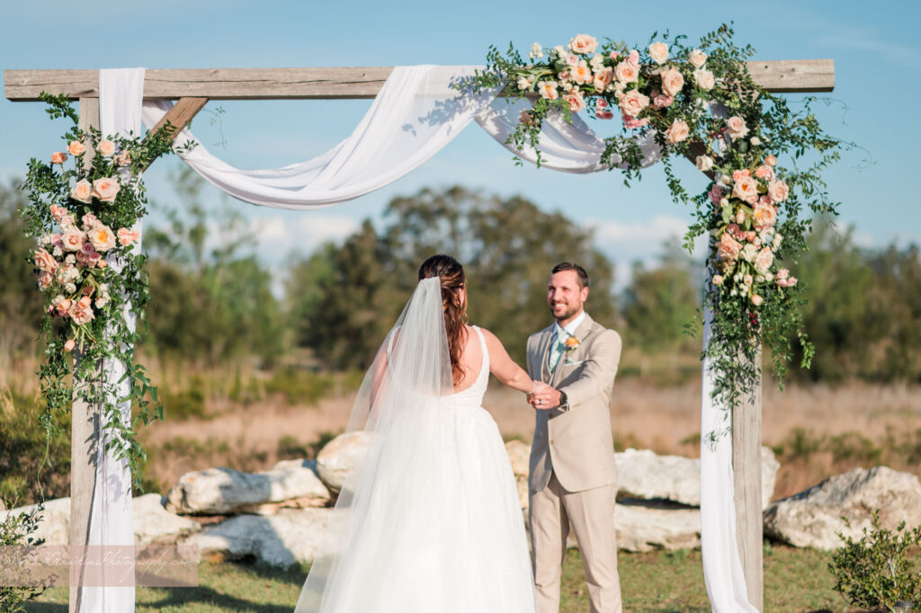 Groom awaits his bride under wedding arch decorated by pink florals and fairy tale vines by Lily's Flower Shop at Acres of Grace Family Farms wedding venue