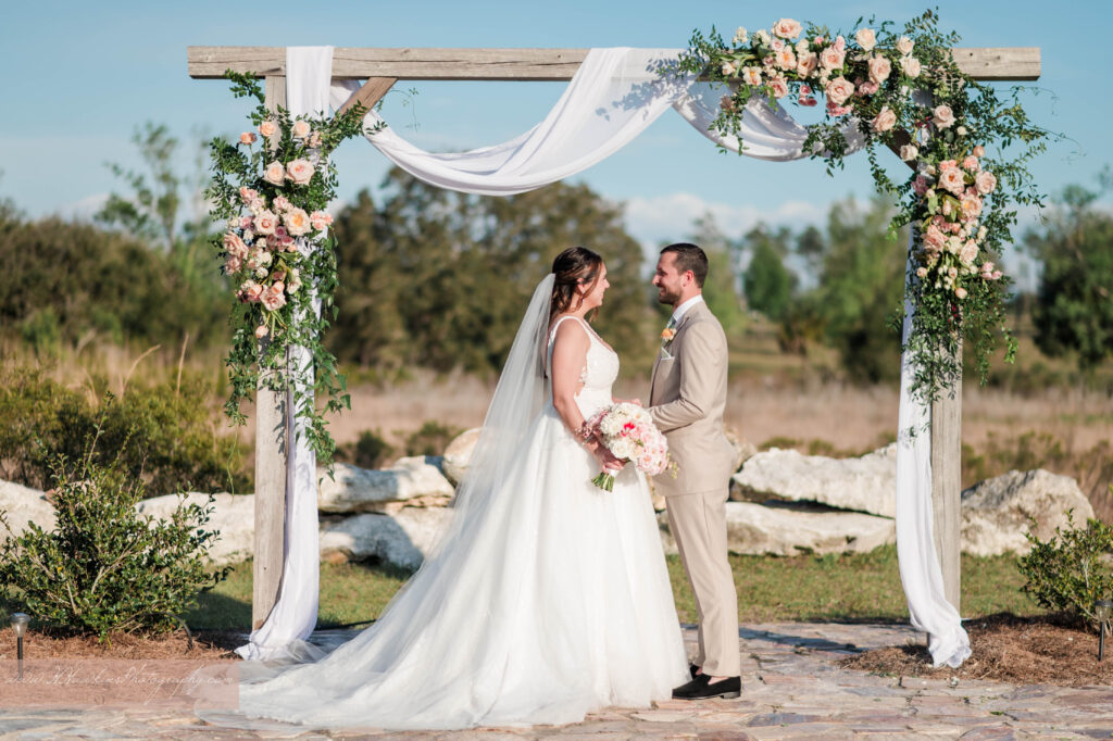 Groom and bride under wedding arch decorated by pink florals and fairy tale vines by Lily's Flower Shop at Acres of Grace Family Farms wedding venue