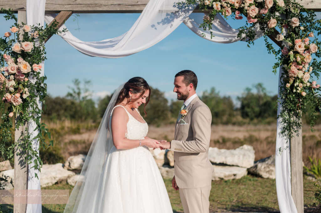 Groom and bride exchange rings under wedding arch decorated by pink florals and fairy tale vines by Lily's Flower Shop at Acres of Grace Family Farms wedding venue