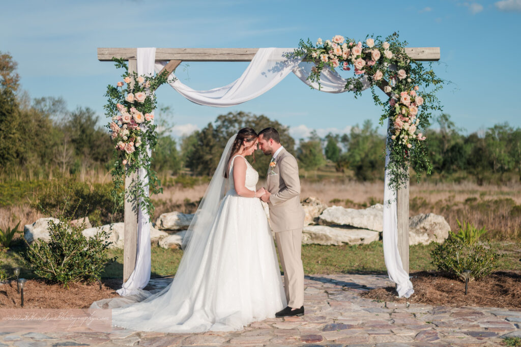 Groom and bride pray together under wedding arch decorated by pink florals and fairy tale vines by Lily's Flower Shop at Acres of Grace Family Farms wedding venue