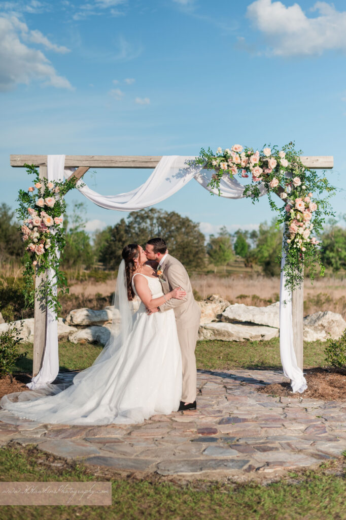Groom and bride kiss under wedding arch decorated by pink florals and fairy tale vines by Lily's Flower Shop at Acres of Grace Family Farms wedding venue