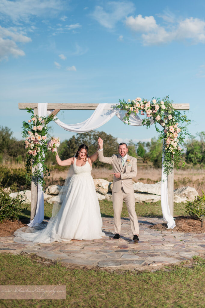Groom and bride celebrate under wedding arch decorated by pink florals and fairy tale vines by Lily's Flower Shop at Acres of Grace Family Farms wedding venue