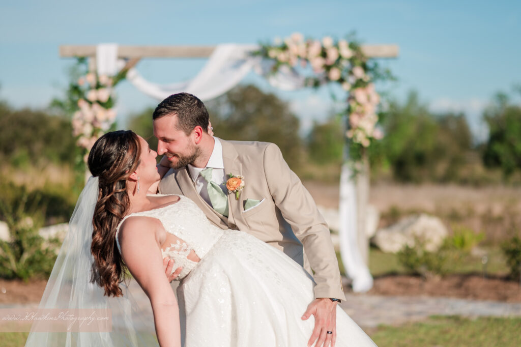 Groom and bride dip kiss under wedding arch decorated by pink florals and fairy tale vines by Lily's Flower Shop at Acres of Grace Family Farms wedding venue