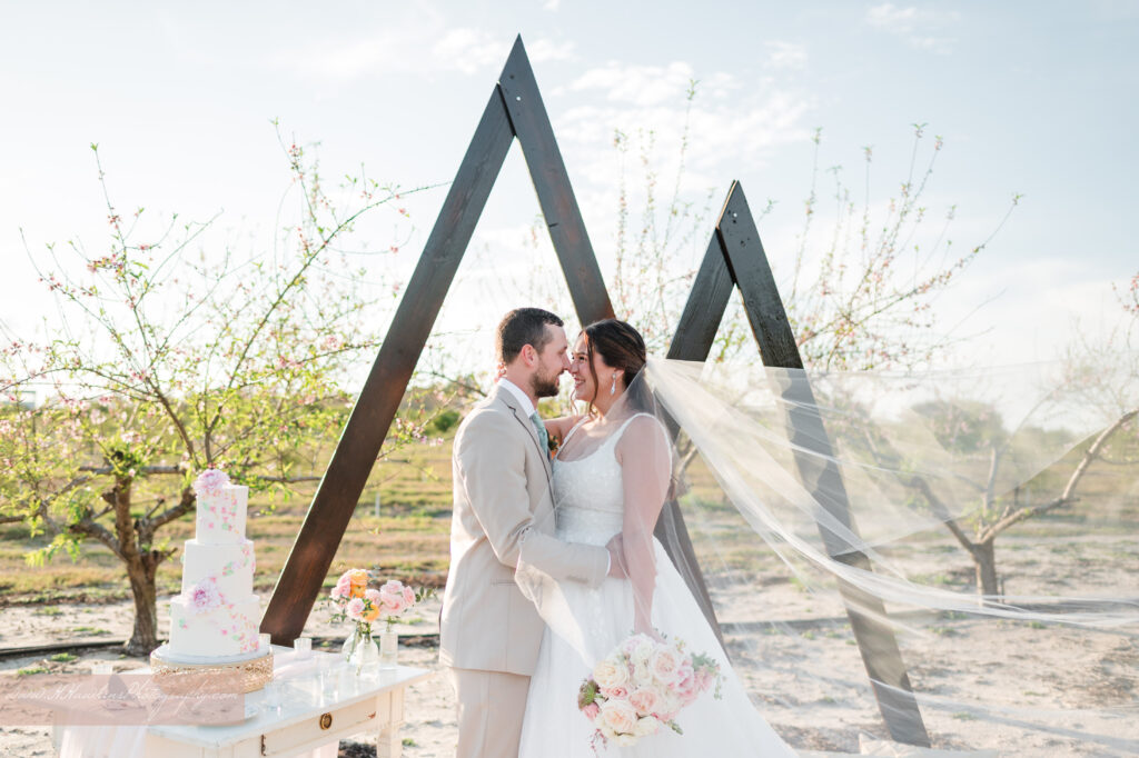Bride and groom kiss in peach blossom orchard at Acres of Grace Family Farms in front of triangle arches with wedding cake