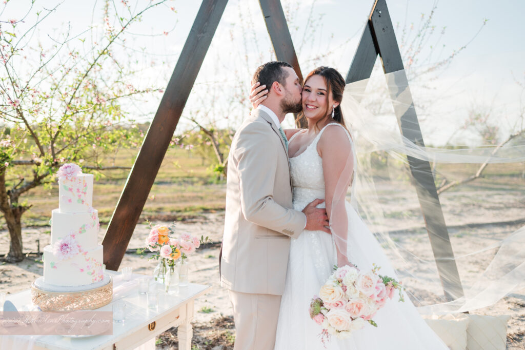 Bride and groom kiss in peach blossom orchard at Acres of Grace Family Farms in front of triangle arches with wedding cake