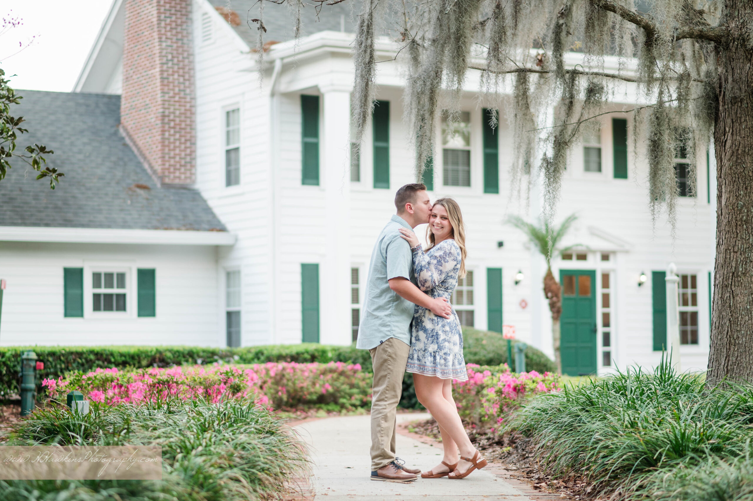 Bride smiles at camera while groom kisses her on the temple in front of Cypress Grove Estate House with spanish moss hanging off the trees