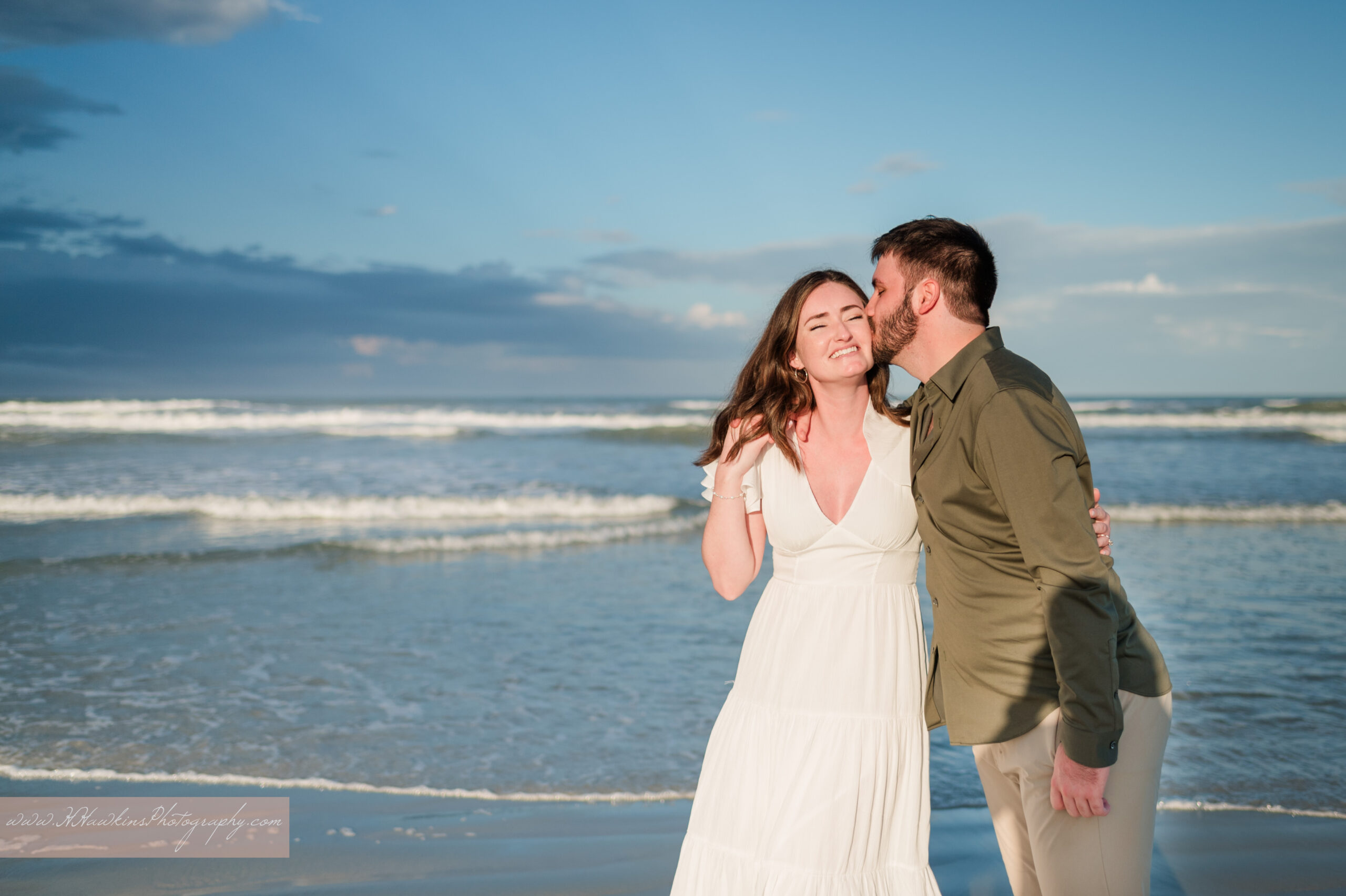 Groom kisses bride to be on the cheek in front of ocean waves at New Smyrna Beach engagement photo session
