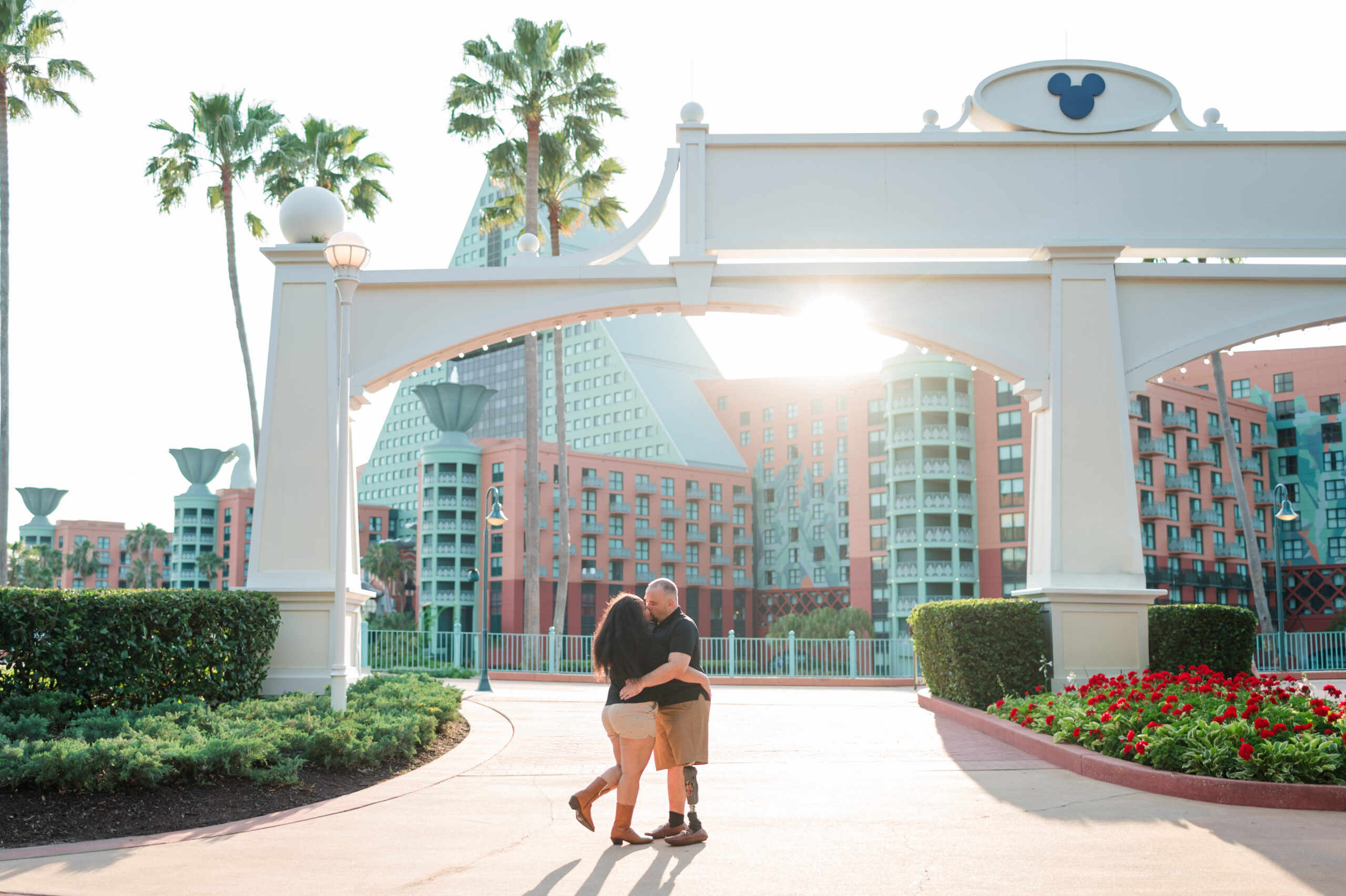 Bride and groom to be stand in front of Disney's Dolphin Resort at the Boardwalk with the sun peeking through the palm trees and Mickey Ear archway