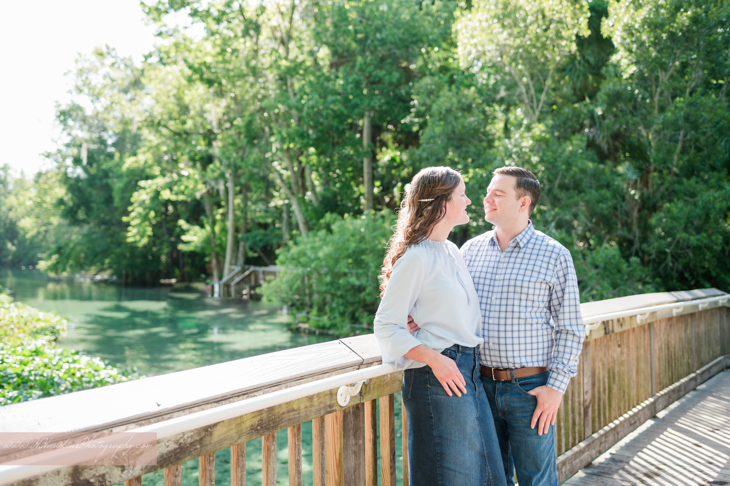 A girl in a jean skirt and guy in button up shirt stand in front of the blue-green waters on a wooden bridge of Wekiwa Springs State Park for their couples engagement photos by Orlando wedding photographer