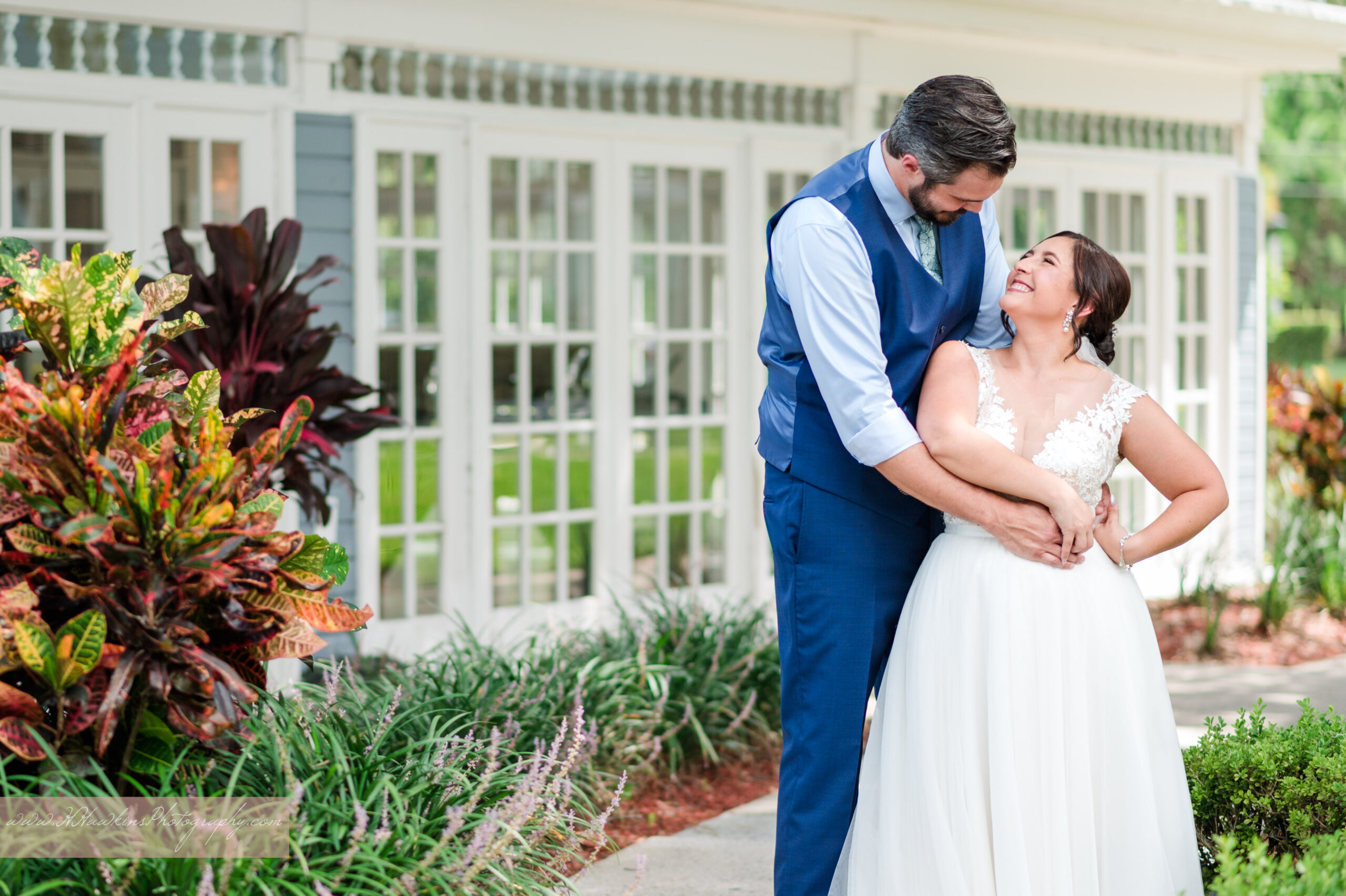 Bride and groom in a navy blue vest smile at each other outside the white and blue Highland Manor garden