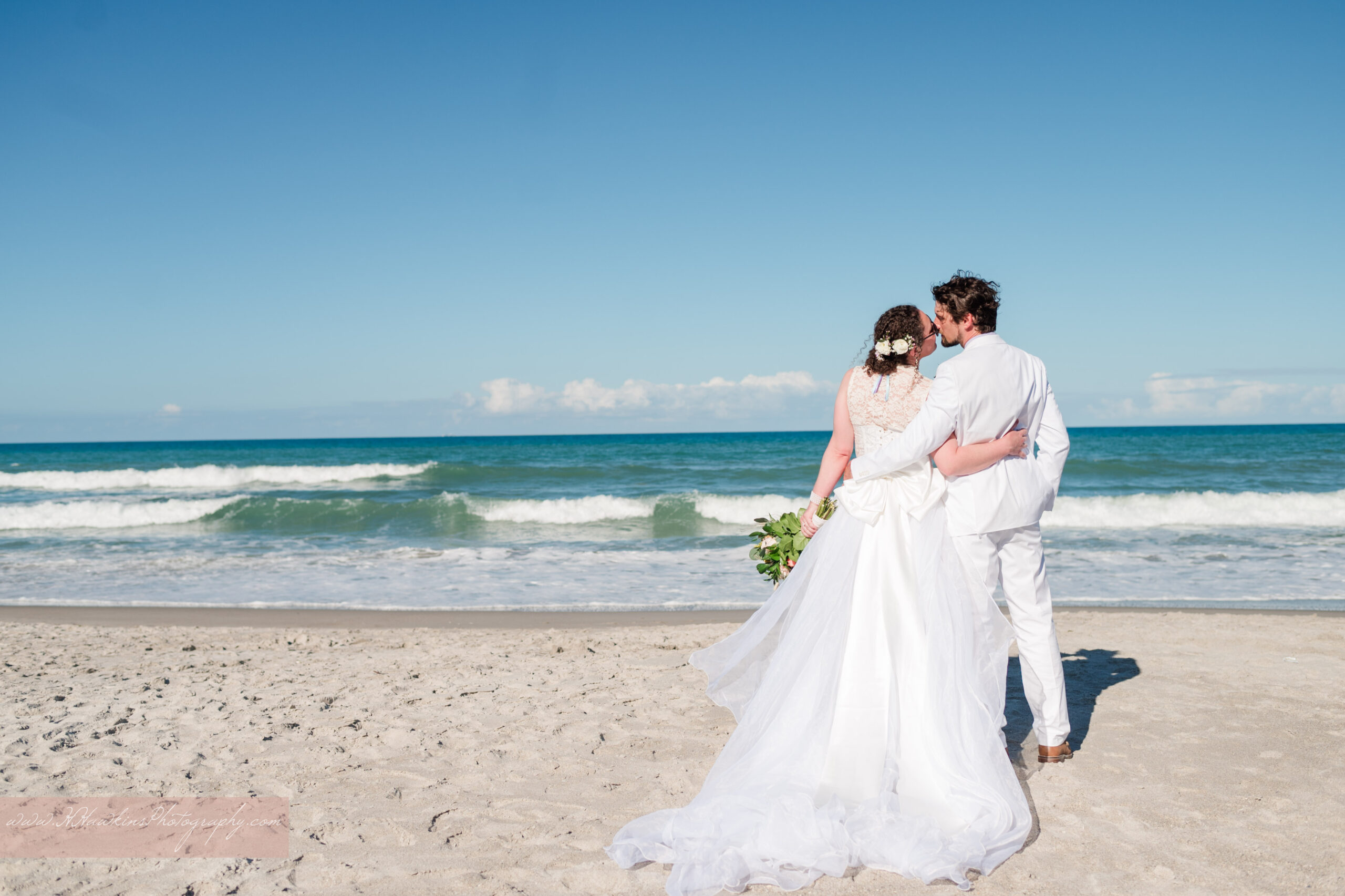 Bride in white dress and groom in white on the beach at The Tides Collocated Club on Patrick Space Force Base in the turquoise ocean behind them