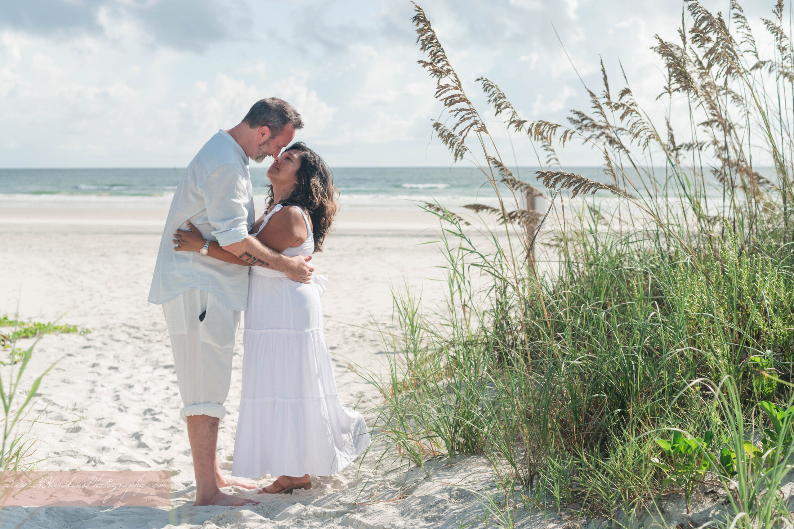 Photos of engaged couple kissing wearing white during their Smyrna Dunes Park engagement session with the sea oats and ocean behind them by New Smyrna Beach Photographer