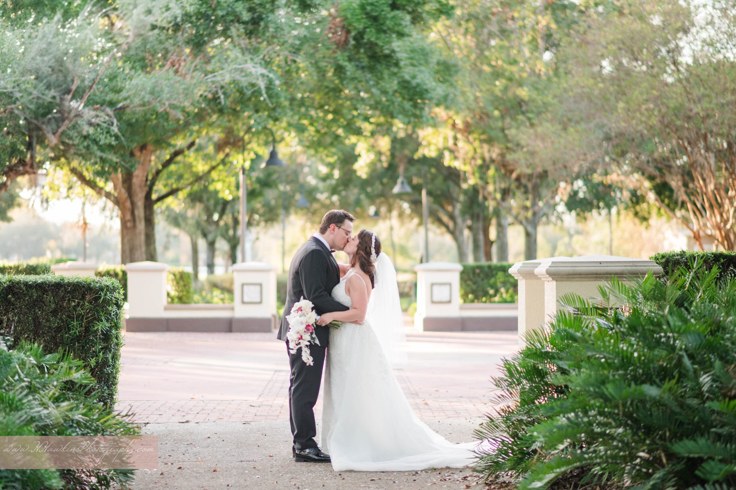 Bride and groom share a kiss beneath a canopy of sunlit trees that forms a natural arch behind them at Harmony Preserve, captured beautifully by an Orlando wedding photographer during their La Bella Rose Ballroom wedding