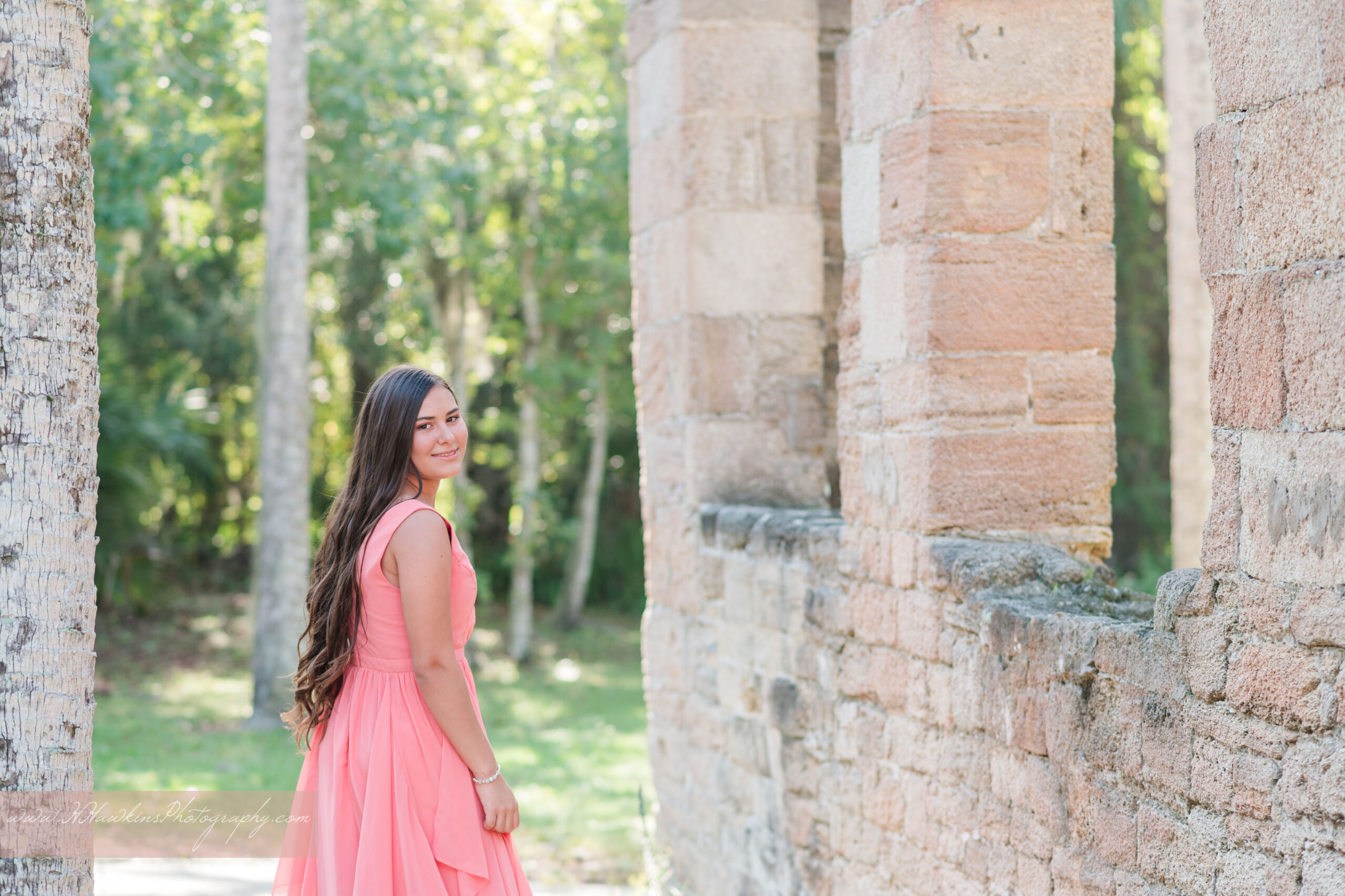 Quinceanera photoshoot of girl in a coral pink flowy gown looking over her shoulder at the historic site of the Sugar Mill Ruins in New Smyrna Beach by Orlando photographer