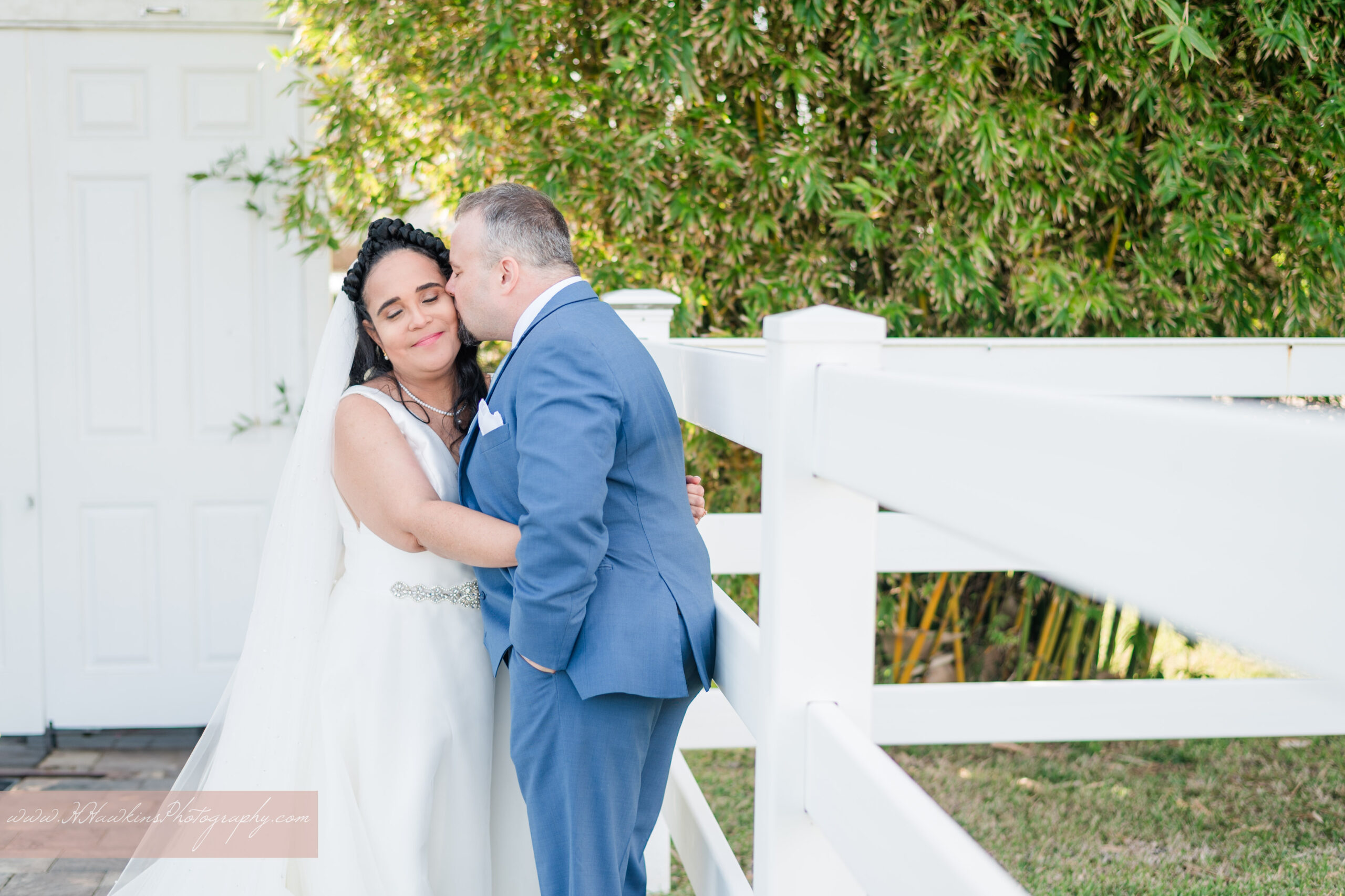 Photo of bride in white dress and long veil and groom in blue suit along the white fence lines at Dragonfly Farms in Palm Bay FL by Orlando wedding photographer